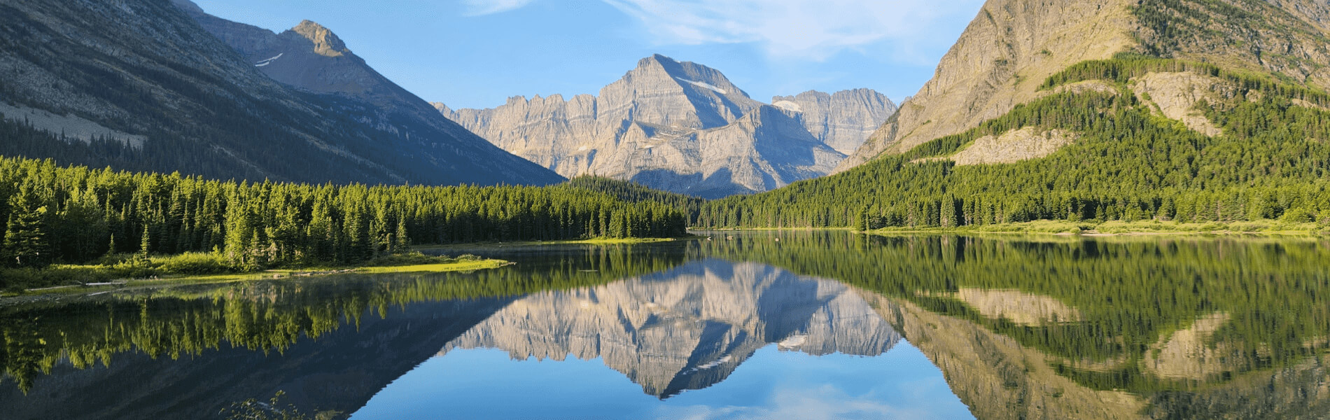 A wide panoramic view of a calm mountain lake reflecting surrounding evergreen forests and towering rocky peaks under a clear blue sky.