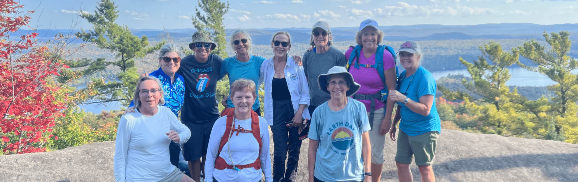 A group of hikers standing together on a rocky overlook surrounded by colorful autumn foliage, with a wide view of forested hills, lakes, and a bright sky in the distance.
